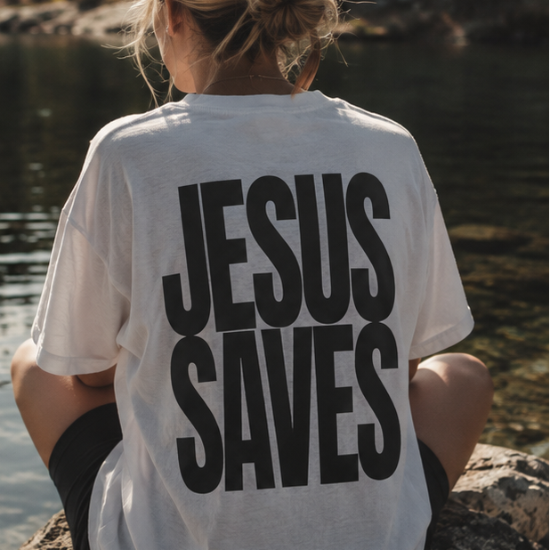 Woman seen from behind wearing white T-shirts with large dark gret/black text reading 'JESUS SAVES' on the back, set against a lakeside forest.