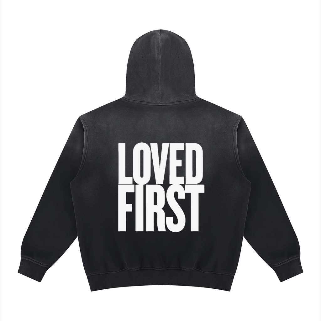 A black hooded sweatshirt shown from the back, featuring the words 'LOVED FIRST' printed in large, bold white block letters across the center.