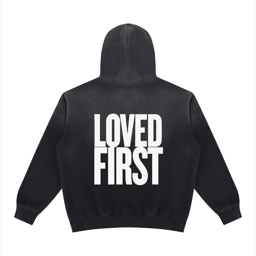 A black hooded sweatshirt shown from the back, featuring the words 'LOVED FIRST' printed in large, bold white block letters across the center.