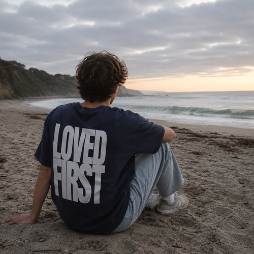 A person sitting on a sandy beach facing the ocean, wearing a navy blue t-shirt with the words 'LOVED FIRST' printed in bold white letters on the back.