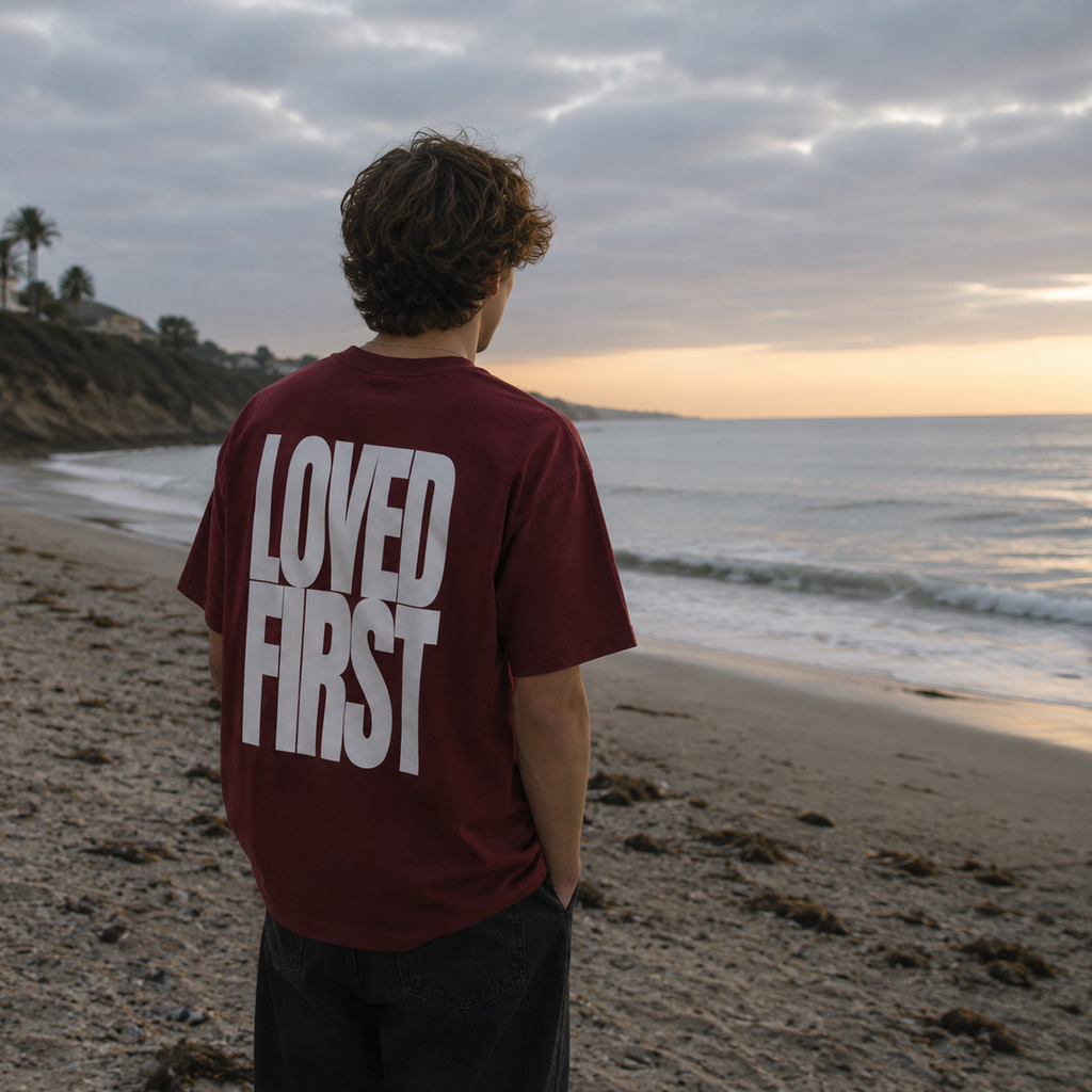 A person standing on a sandy beach at sunset, viewed from behind, wearing a Wine Red t-shirt with the words 'LOVED FIRST' printed in large white letters on the back.