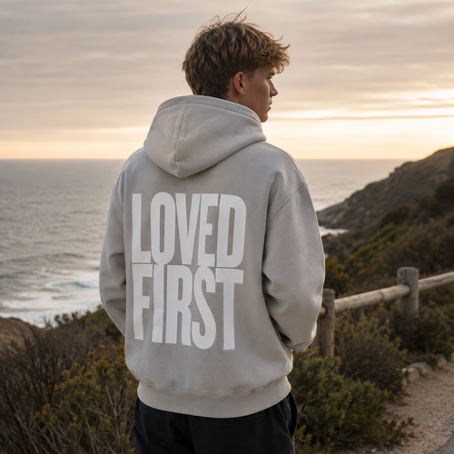 A man standing outdoors by the ocean at sunset, viewed from behind, wearing a light gray hoodie with the words 'LOVED FIRST' printed in large white letters on the back.