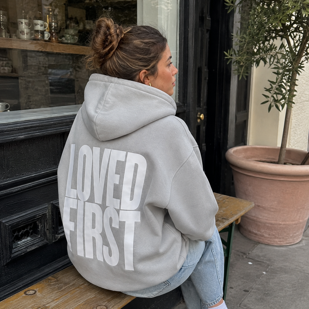 A woman sitting on a wooden bench outdoors, wearing a light gray oversized hoodie with the words 'LOVED FIRST' printed in large white letters across the back. She is also wearing light blue jeans, white socks, and gray sneakers.