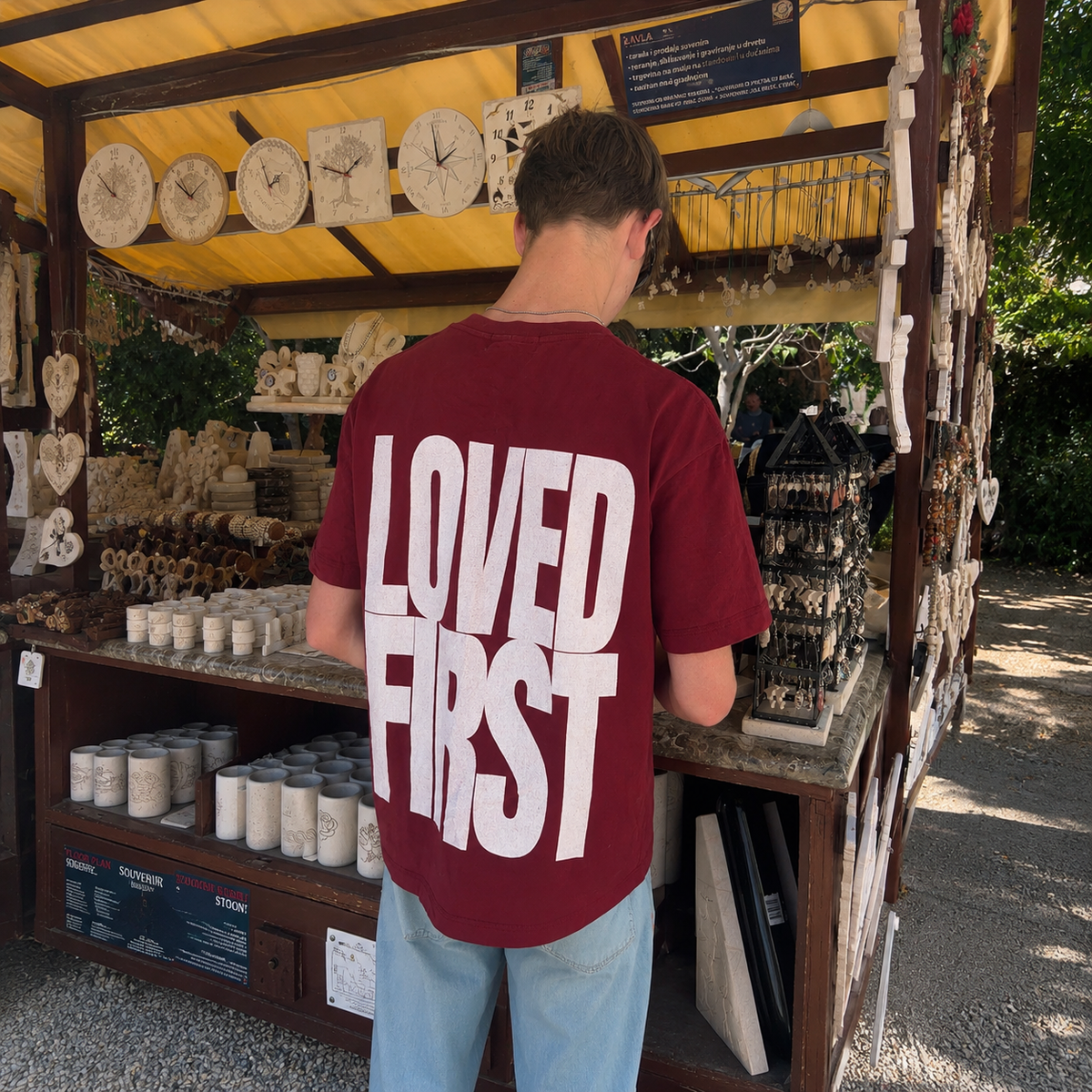 A man viewed from behind wearing a wine red t-shirt with 'LOVED FIRST' printed in bold white letters, standing at a wooden market stall filled with handmade items.