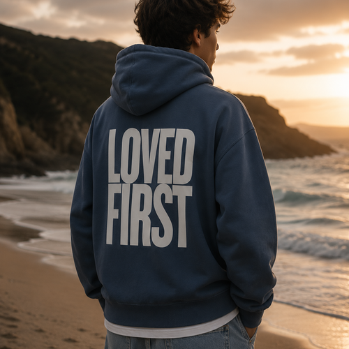 A person standing on a beach at sunset, viewed from behind, wearing a washed blue hoodie with large white text reading 'LOVED FIRST' on the back and light blue jeans.