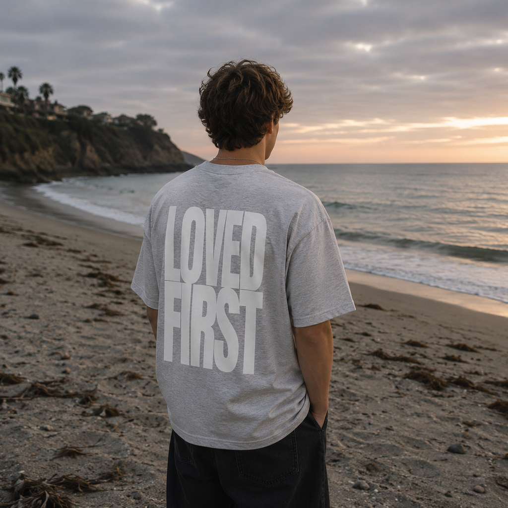 A man standing on a beach with his back to the camera, wearing a light gray t-shirt with the words 'LOVED FIRST' printed in large white letters on the back, and dark pants. The beach, ocean, and a cloudy sunset sky are visible in the background.