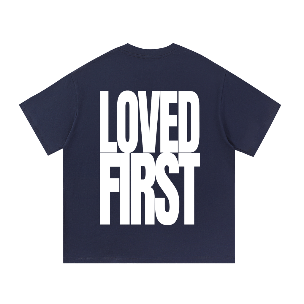 A navy blue short-sleeve t-shirt with the words 'LOVED FIRST' printed in large, bold white block letters across the front, shown on a plain white background.