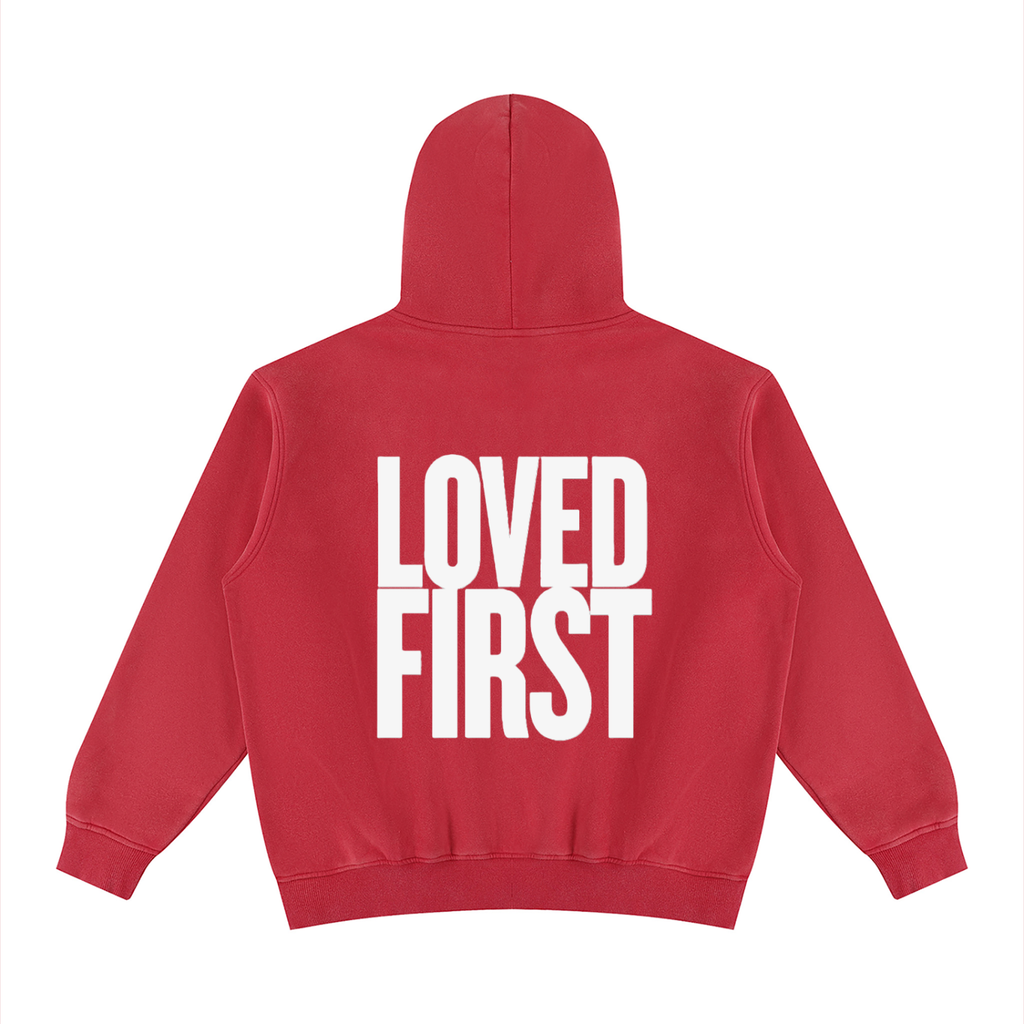 A red pullover hoodie shown from the back, featuring the words 'LOVED FIRST' printed in large, bold white block letters across the center.