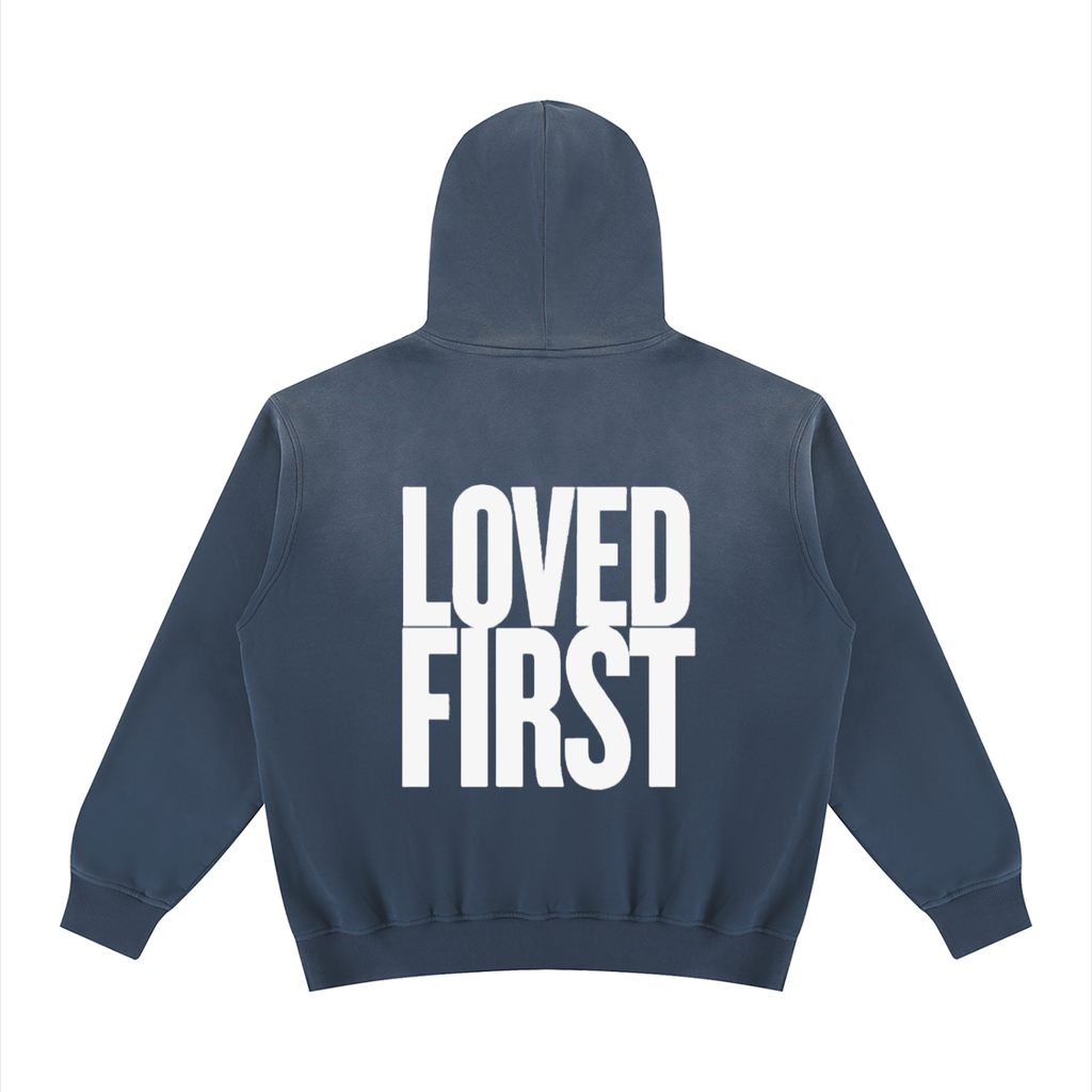 A black hooded sweatshirt shown from the back, featuring the words 'LOVED FIRST' printed in large, bold white block letters across the center.