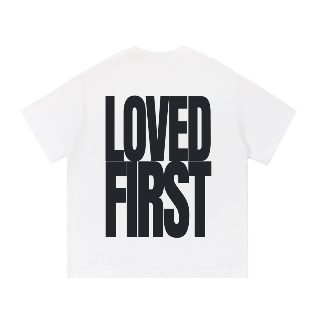 A white short-sleeve t-shirt shown from the back, featuring the words 'LOVED FIRST' printed in a large, bold, black sans-serif font.