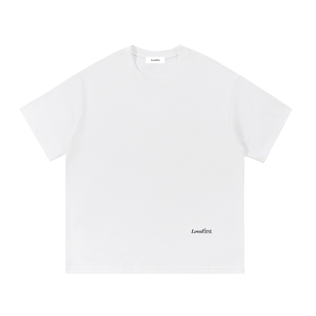 A white short-sleeve crewneck t-shirt with a small black 'Lovedfirst' logo printed on the lower right front, displayed flat against a solid white background.
