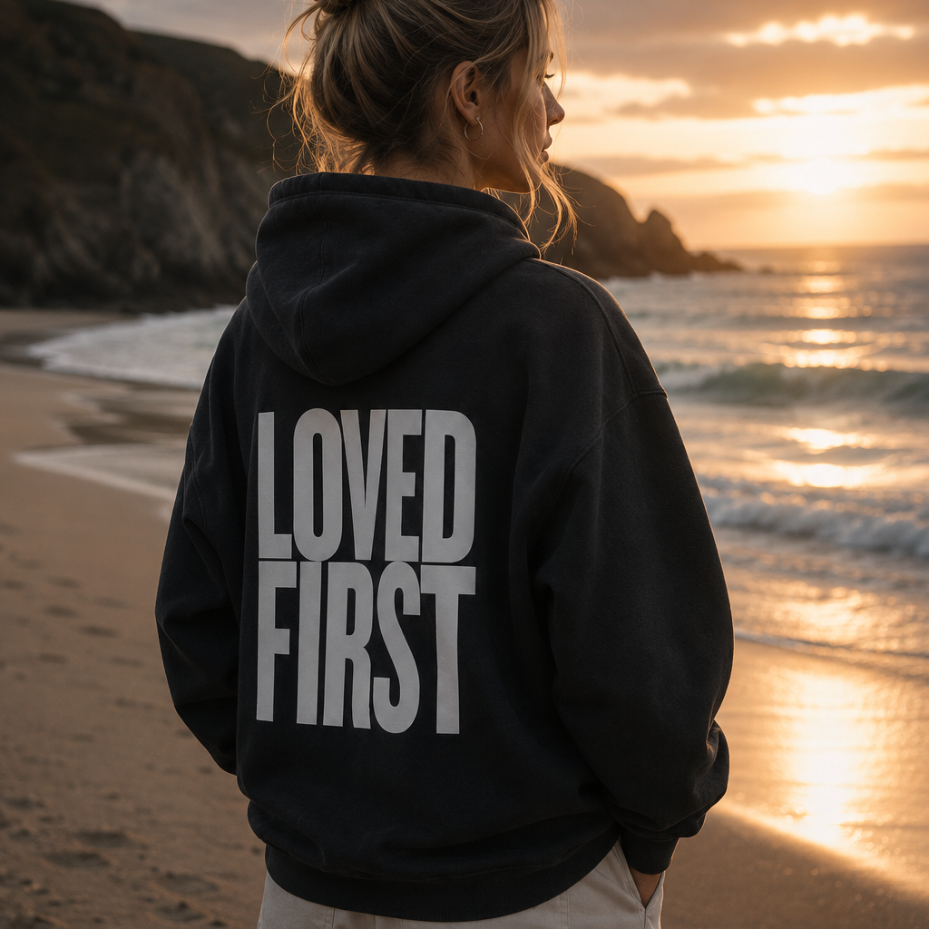A woman stands on a beach at sunset facing the ocean, wearing a black hoodie with 'LOVED FIRST' printed in bold white letters on the back.