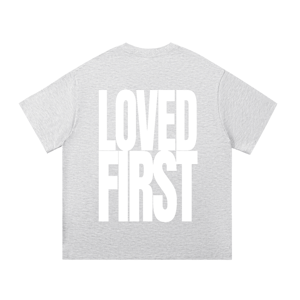 A light heather gray short-sleeve t-shirt with the words 'LOVED FIRST' printed in large, bold white block letters across the back.
