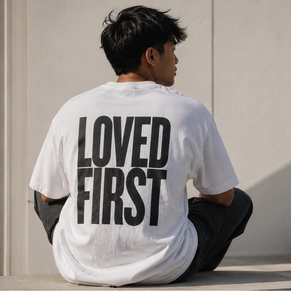 A man sitting outdoors on a concrete ledge, viewed from behind, wearing a white oversized T-shirt with the words 'LOVED FIRST' printed in bold black letters on the back.