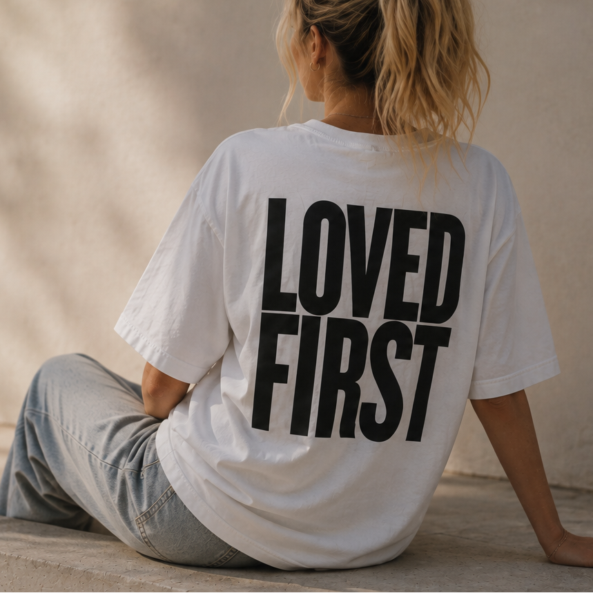 A woman sitting on a concrete ledge outdoors, viewed from behind, wearing a white oversized T-shirt with the words 'LOVED FIRST' printed in bold black letters on the back.